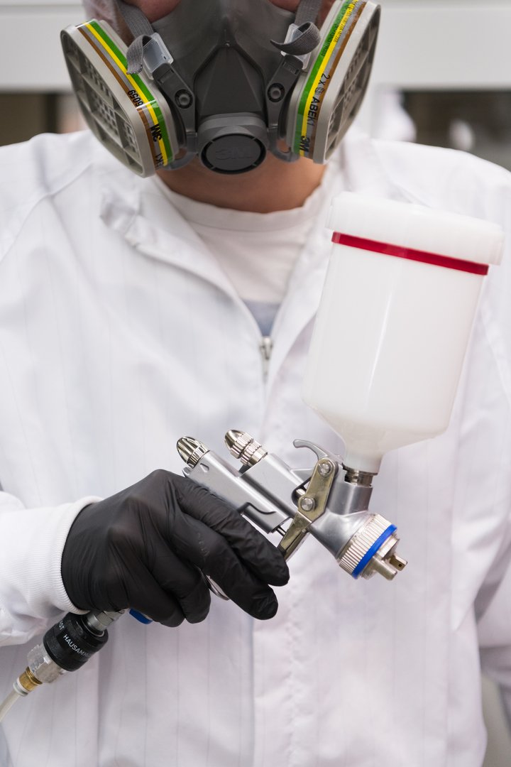 A technician wearing a protective mask and gloves prepares to apply lacquer using an airbrush