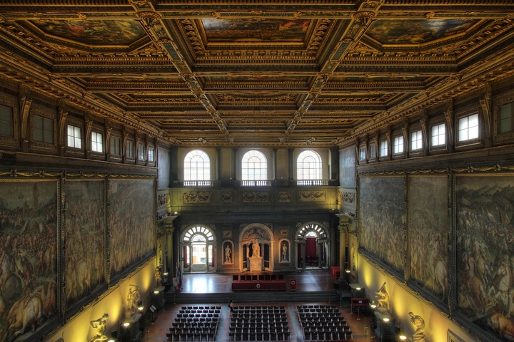 The Salone dei Cinquecento in the Palazzo Vecchio: 54 metres long, 23 wide and 18 high. An immense coffered ceiling rises above the dinner tables, while the walls bear monumental compositions created under Giorgio Vasari's direction for Cosimo I de' Medici, glorifying Florence, its conquests and ducal power.